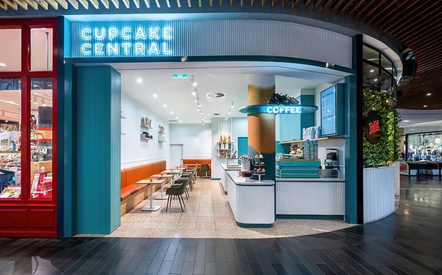 Interior view of a brightly lit cupcake and coffee shop kiosk in a retail or commercial setting. The compact café features a curved teal and white service counter, integrated display shelving, orange upholstered banquette seating, small tables, patterned tile flooring, red-framed glazed frontage, and prominent neon “CUPCAKE CENTRAL” and “COFFEE” signage. The space has a playful, polished, contemporary hospitality fit-out.