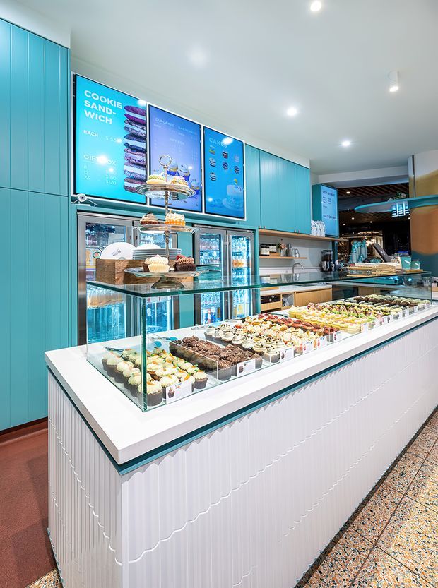 Bright contemporary dessert and gelato shop interior with a long white fluted service counter, glass display cases filled with pastries and desserts, turquoise built-in cabinetry, illuminated drinks refrigerators, menu boards, and polished terrazzo-style flooring. The space has a clean, colourful, highly finished commercial fit-out.
