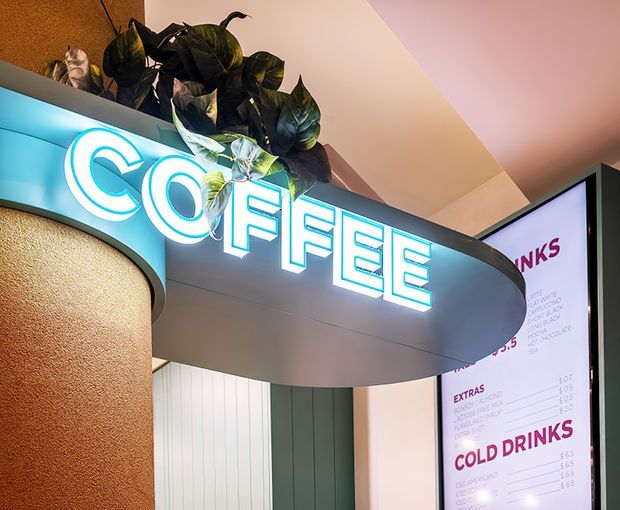 Close-up interior view of a stylised café counter or kiosk, dominated by a glowing cyan-white “COFFEE” sign mounted on a curved dark-edged canopy. The space features a textured warm brown curved wall, pale pink and green vertical wall panels, an illuminated menu board, and decorative trailing foliage above the signage. The image has a contemporary commercial hospitality aesthetic with bold neon lighting and rounded forms.