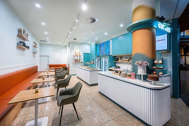 Bright contemporary commercial café or dessert shop interior with a long orange upholstered banquette, small timber tables, dark upholstered chairs, white fluted service counters, turquoise cabinetry and wall panels, terrazzo flooring, and prominent cylindrical column detailing. Refrigerated display cases, shelving, and counter displays are visible throughout the open customer area.