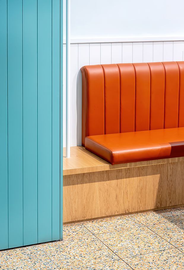 A brightly lit contemporary interior detail featuring a built-in orange upholstered banquette with vertically channelled backrest, set against white vertically panelled walls. The seating is integrated over a timber-clad base with a light timber ledge, beside a tall turquoise fluted panel or door. The floor is finished in colourful terrazzo-like tiles with visible grout lines.