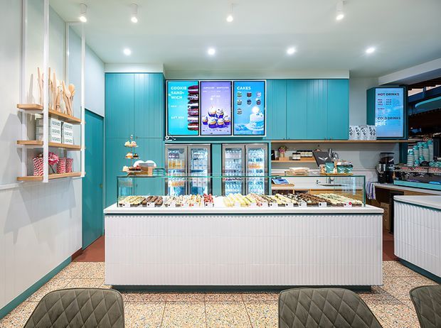 A brightly lit contemporary commercial bakery or dessert shop interior, viewed from the customer seating area toward a central white fluted display counter. Turquoise-blue full-height cabinetry and wall panels frame illuminated menu screens, refrigerated display cases, shelving, and a service counter. Terrazzo-style flooring, pale walls, recessed spotlights, and dark quilted seating complete the polished fit-out.