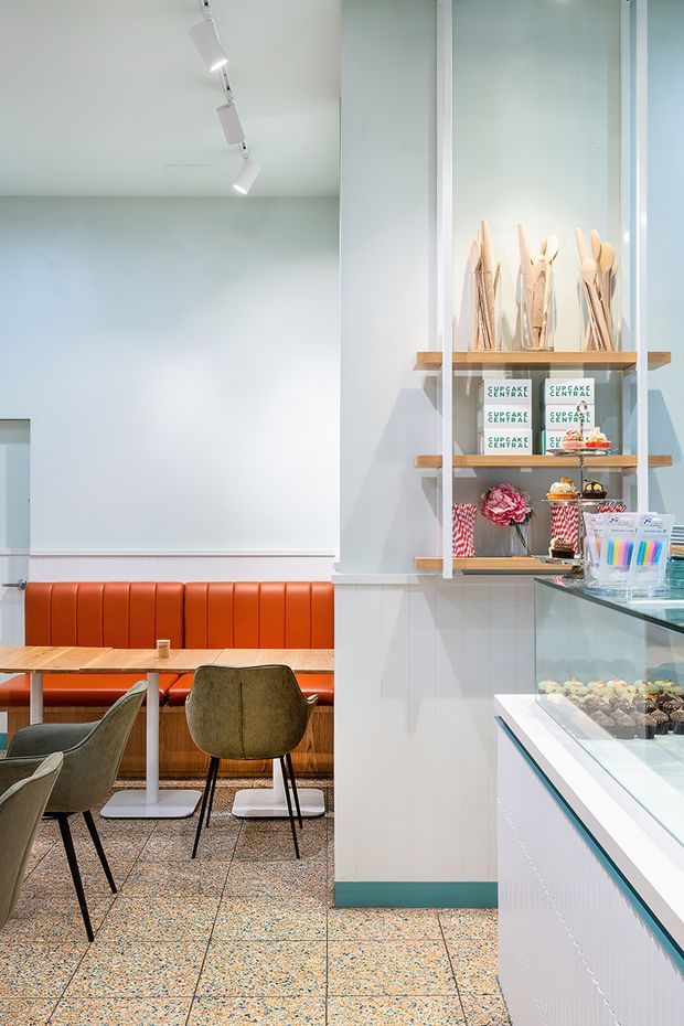 Bright contemporary cupcake shop and café interior with a compact dining area, orange upholstered banquette, wooden tables, upholstered chairs, terrazzo-style flooring, and a glass-fronted pastry display counter. White shelving displays cupcake boxes and decorative utensils, while track lighting and pale mint-blue walls create a clean, playful retail setting.