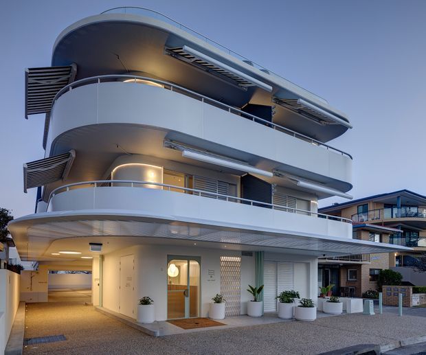 Exterior view of a contemporary multi-storey residential apartment building at dusk. The building features rounded projecting balconies, curved slab edges, white rendered walls, horizontal metal balustrades, integrated exterior lighting, louvred sun-shading panels, and a broad flat roof form. Ground level includes a glazed entry door, recessed pedestrian entry, potted plants, and a covered driveway or access passage. A neighbouring apartment building is visible in the background.