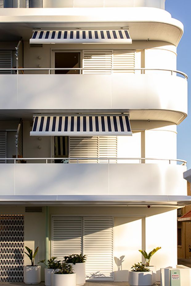 Exterior view of a multi-storey residential building with rounded balcony edges, white rendered walls, metal balustrades, external roller shutters and striped retractable awnings. Potted plants line the ground-level frontage, and strong sunlight emphasizes the clean curved facade.