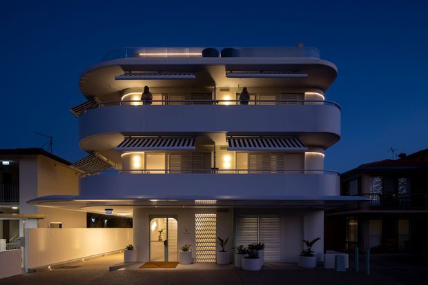 Twilight exterior photograph of a compact multi-storey residential building with strongly rounded balconies, curved projecting rooflines, horizontal metal railings, striped retractable awnings, warm interior lighting, and a recessed ground-floor entry. Adjacent buildings and potted plants frame the property.