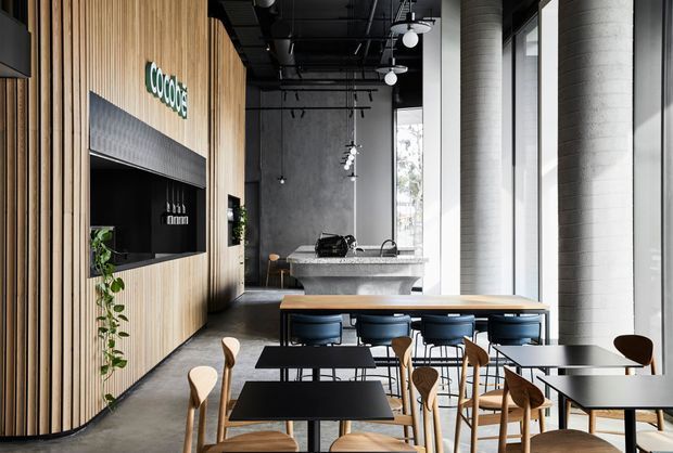 A contemporary commercial café interior with a long communal timber table and black café tables in the foreground, surrounded by light timber chairs. The space features a vertical timber slat feature wall with a built-in service or display opening and signage, a dark exposed ceiling with suspended pendant lighting, polished concrete floors, a concrete service counter, and tall floor-to-ceiling windows. The palette is predominantly black, grey, light wood, and white, creating a refined industrial atmosphere.