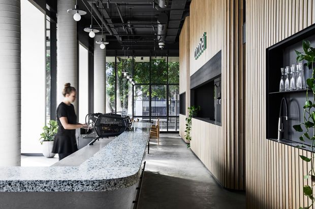A contemporary commercial café or hospitality interior with a long speckled stone service counter, integrated coffee machine, black open ceiling with exposed services, tall glazing, and a feature wall clad in vertical light timber battens. Open shelving displays glassware, while pendant lights, plants, and timber seating add detail. The space is bright, linear, and refined with an industrial-modern fit-out.