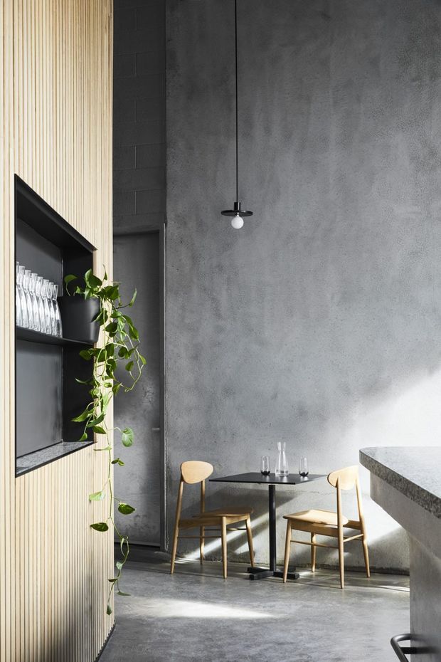 A compact, high-ceilinged contemporary café or hospitality interior with a small dining table and two light timber chairs beside a textured grey concrete wall. A black built-in shelving and service/display unit is integrated into a vertical light timber slat wall, with glassware and a trailing plant. A pale stone or concrete counter occupies the right foreground, while a minimalist black pendant hangs above the table. Strong sunlight creates bright patches and shadows across the grey concrete floor and walls.