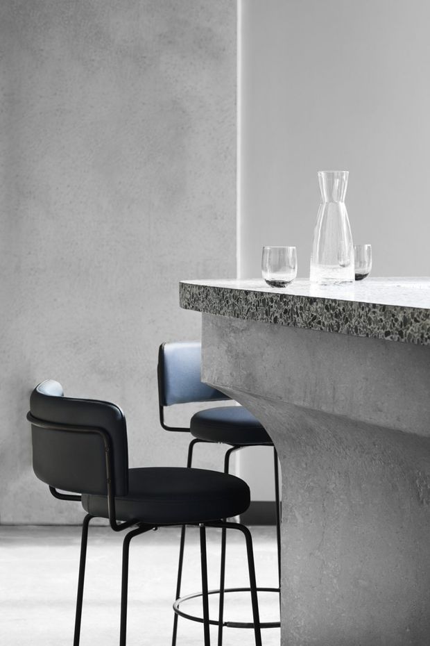 Monochrome minimalist interior showing a sculptural concrete kitchen or bar island with a heavily textured dark stone benchtop. Three black upholstered stools sit beneath the overhanging counter, with clear glassware arranged on top. The space has smooth grey plaster or concrete walls and flooring, creating a restrained, industrial atmosphere.