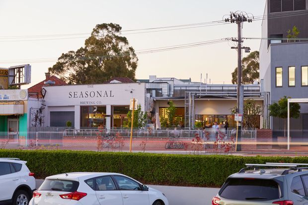 Street-level exterior view of a contemporary mixed-use urban streetscape, centred on The Seasonal Brewing Co. building with outdoor dining or forecourt activity, adjacent modern commercial buildings, utility infrastructure, planted street-edge hedges, and blurred traffic in the foreground.