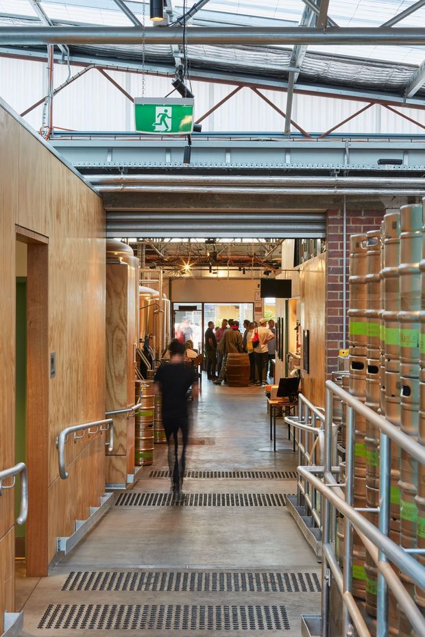 A view down an industrial brewery or production venue corridor, with concrete flooring, grated anti-slip stair treads, stainless-steel handrails, large stainless-steel brewing vessels and kegs, exposed services, steel roof structure, translucent roof panels, timber-lined walls, and exposed brick. People gather in a taproom or public area in the distance. The space combines robust industrial infrastructure with warm timber and brick finishes.