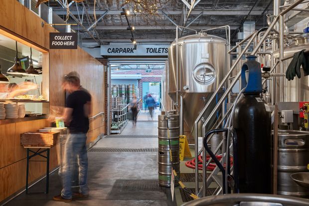 Interior view of a brewery taproom or beer hall with an industrial production area integrated into the customer space. A timber-clad food service counter occupies the left side, while stainless-steel brewing tanks, pipes, kegs, and equipment line the right. Industrial exposed ceiling services, steel framing, concrete flooring, overhead wayfinding signs, and a visible passage toward the toilets and carpark reinforce the utilitarian hospitality setting.
