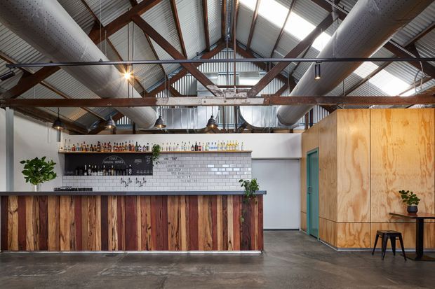A spacious industrial-style bar or hospitality interior beneath a high pitched corrugated-metal roof. Exposed timber roof trusses, large silver ventilation ducts, pendant lights, and suspended services create a strong warehouse character. The bar front is clad in vertically arranged reclaimed timber boards, with a white subway-tile backsplash, dark shelving stocked with bottles, and a plywood-clad service volume to the right. Polished concrete flooring and muted teal door accents complete the utilitarian yet warm fit-out.