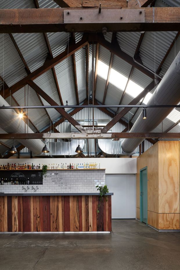 Interior view of a rustic-industrial bar or hospitality venue. The space features a high pitched corrugated metal roof with exposed timber trusses, exposed ductwork, suspended track lighting and pendant lights, a white subway-tile bar backsplash, reclaimed timber bar front, polished concrete flooring, and plywood-clad cabinetry or wall volumes. Bottles and a chalkboard menu line the bar.