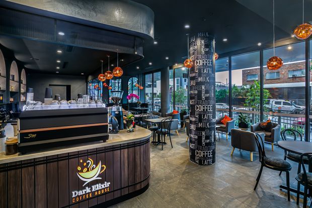 A stylish urban coffee shop interior with a curved dark timber service counter, commercial espresso machine, illuminated cafe branding, high black ceilings with recessed lighting, pendant globe lights, patterned feature column, and extensive floor-to-ceiling glazing. Seating includes small round tables, black chairs, and lounge armchairs overlooking the street.