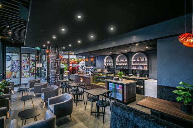 A contemporary café interior with a dark ceiling, polished concrete-style flooring, a long service counter and display cases, illuminated beverage shelving, café tables, black metal chairs, upholstered lounge seating, decorative pendant lights, and colourful glazed artwork near the entrance.