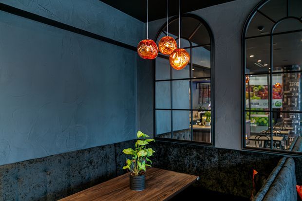Moody commercial interior featuring a dark textured plaster wall, black-framed arched industrial-style windows, black stone-look lower wall finish, a timber dining table with potted plant, and three glowing amber pendant lights. Seating and a café or restaurant interior are visible through the windows and reflections.