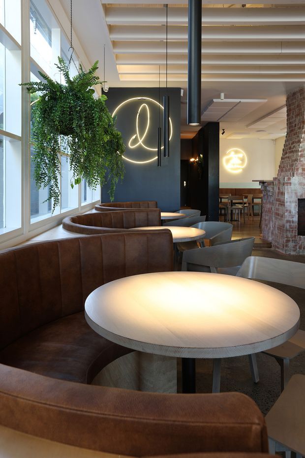 A stylish commercial hospitality interior, likely a café or restaurant, with curved brown leather banquette seating arranged around round tables. The space features large industrial-style glazing, exposed brickwork, dark painted feature walls, suspended linear and cylindrical pendant lighting, neon wall signage, and a ceiling with exposed timber battens and services. Hanging greenery softens the otherwise contemporary industrial fit-out.