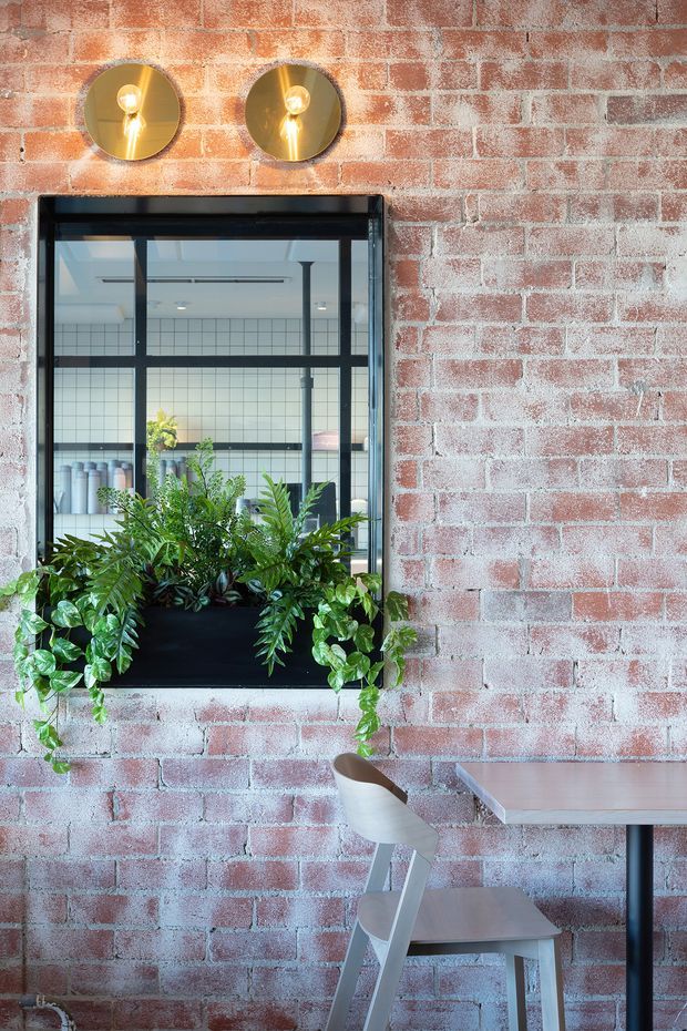 A compact industrial-style interior with an exposed red brick wall, black-framed internal window, and a black planter box filled with lush trailing greenery. Two round brass wall lights illuminate the brickwork above the window. A small light-toned table and chair occupy the lower right, while the window reveals a tiled workspace or adjoining area beyond.
