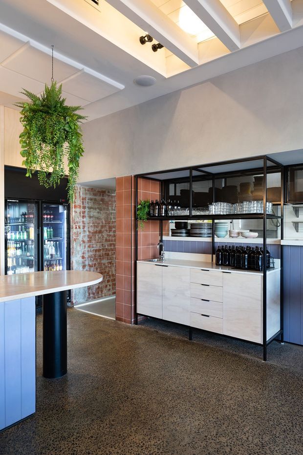 A bright, contemporary commercial hospitality interior, likely a café, bar, or restaurant service area. The space features black-framed glazed shelving and cabinetry, a pale stone or solid-surface benchtop, dark blue lower cabinetry, terracotta rectangular tile, exposed brick, and a polished aggregate floor. Bottles, glassware, and crockery are displayed in the open shelving. A large hanging plant and round communal table add visual interest. Exposed white ceiling beams and a rooflight or clerestory bring in natural light.