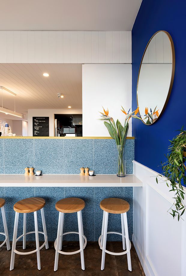 Bright, compact café-style dining counter with three white stools and round timber seats in front of a blue textured tiled or speckled feature wall. A white benchtop runs across the counter, with small condiment settings and a vase of bird-of-paradise flowers. The space has a vivid cobalt-blue wall, white vertical wall panelling and ceiling lining, an oval timber-framed mirror, white dado panelling, dark timber flooring, and a glimpse of a café service area beyond.