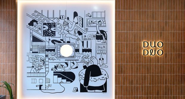 A stylised interior wall feature with a large backlit black-and-white illustrated artwork mounted against a narrow vertical timber-slat wall. To the right, a glowing “DUO” sign is integrated into the slatted wall. A small plant is partially visible at the lower left.