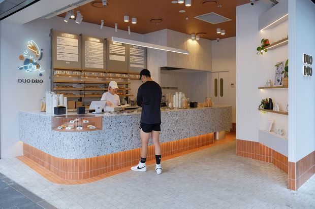A bright contemporary dessert or bakery shop interior with a terrazzo-clad service counter, orange ceramic tile base, warm wood ceiling, menu boards, display shelving, and integrated product shelving. Staff and a customer occupy the foreground, while the open counter and back-of-house preparation area define the space.