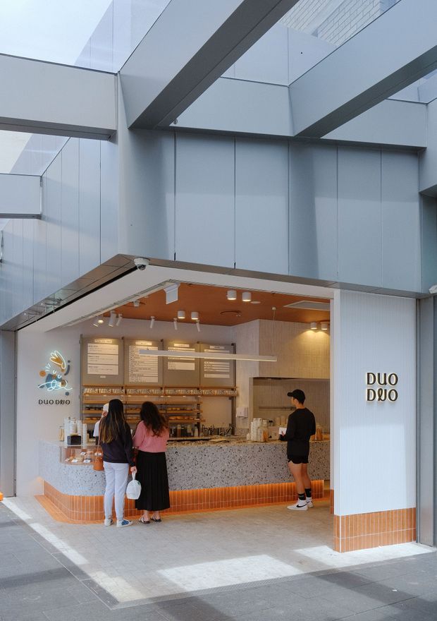 Street-level view of a contemporary café or dessert shop integrated into a modern glazed building. The open storefront features a terrazzo-style service counter, orange ceramic tile base, warm timber-toned ceiling, menu boards, display shelving, pendant and track lighting, and branded wall signage. Large structural beams, glass panels, and pale blue-grey cladding surround the retail frontage.