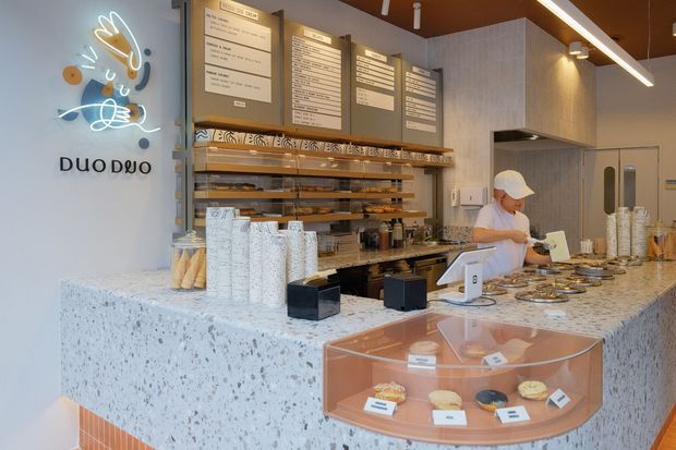 A contemporary café or bakery counter interior with a terrazzo-clad service counter, pastry display case, timber shelving, menu boards, recessed display lighting, and a staff member serving customers. The space features a warm timber ceiling, light grey cabinetry and walls, and polished food-service fixtures.