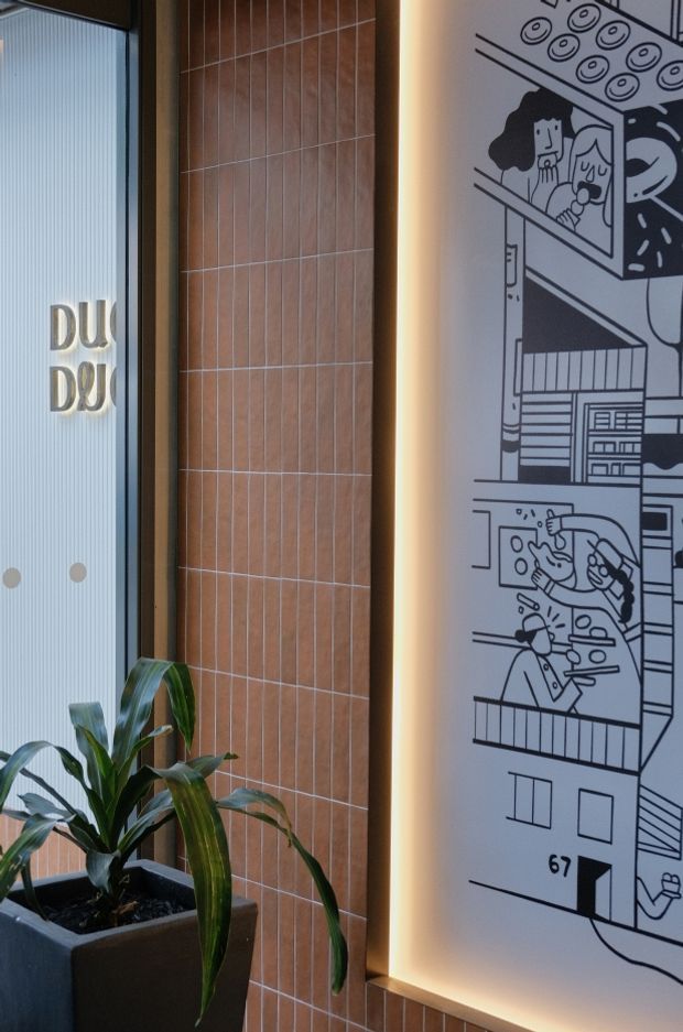 A close-up of a stylised commercial interior feature wall. A tall backlit monochrome illustrated panel depicts people and architectural scenes, bordered by warm timber-toned vertical wall tiles. To the left is a ribbed glass door or window with partial lettering, and a potted indoor plant sits in the foreground.