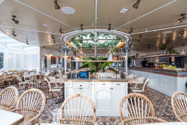 A bright, ornate café or restaurant interior with extensive rattan dining chairs and tables, patterned floor tiles, white cabinetry, a central service island, and a polished bar counter. The space features a glazed or open garden-facing elevation, decorative arches, abundant plants, and ceiling-mounted track lighting.