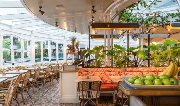 Bright, ornate conservatory-style restaurant interior with white-framed arched glazing, a translucent roof, patterned floor tiles, rattan dining chairs, upholstered banquettes, abundant tropical planting, and suspended warm lighting. The space combines a formal dining area with a lounge-like booth and a prominent fruit display.