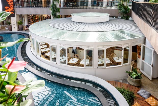Interior view of a bright, multi-level commercial atrium featuring a distinctive circular restaurant or café pavilion with white-framed glazing, a translucent radial roof, and a central raised roof structure. Curved turquoise water features surround the pavilion, with lush indoor planting, balconies, and seating visible around the perimeter.