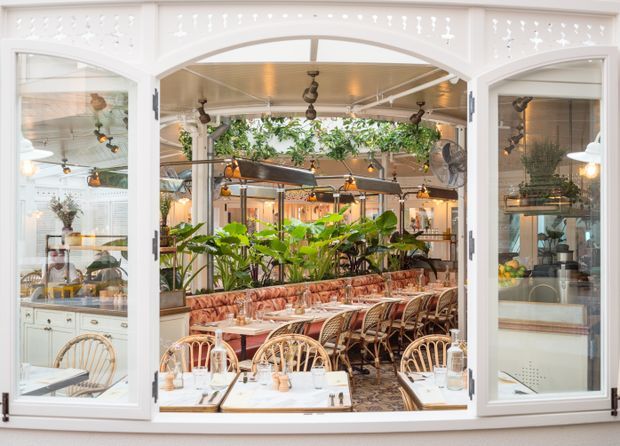 View through open white-framed arched glazing into a bright, plant-filled restaurant or café dining area. The space features rows of marble-topped dining tables, rattan chairs, patterned banquette seating, hanging and wall-mounted lighting, abundant greenery, and a warm decorative ceiling with a glass-roofed or courtyard-like feel.