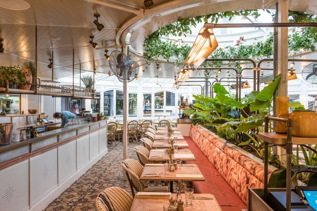 A bright, ornate restaurant interior with a long service counter and open kitchen on the left, closely arranged dining tables and wicker chairs through the centre, and a long patterned banquette on the right. The space features a glazed roof or atrium, abundant indoor planting, decorative pendant lights, exposed metal pipework, ceiling fans, and a warm timber-toned ceiling with detailed traditional architectural elements.