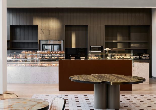 A sophisticated commercial café or bakery interior with a central dark brown service island, illuminated pastry display counters, integrated dark timber cabinetry, appliances, and pale stone finishes. A round stone-topped pedestal table and patterned tiled floor occupy the foreground. The space has a warm, refined, contemporary character with strong built-in joinery and layered lighting.