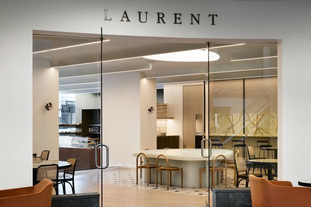 A refined contemporary hospitality or retail interior viewed through full-height glass doors, with a central light stone counter, pale timber wall cabinetry, marble-patterned feature panels, café-style tables and chairs, and layered linear and circular ceiling lighting. The space has warm neutral finishes and a polished, upscale atmosphere. The word “LAURENT” is displayed above the entrance.