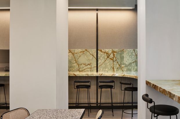 A refined contemporary kitchen and bar area viewed through a partial partition. The space features a marble-look stone backsplash and benchtops with dramatic veining, warm textured wall panels, dark metal bar stools, integrated linear lighting, and pale neutral walls. A small portion of dining furniture and a second benchtop are visible in the foreground.