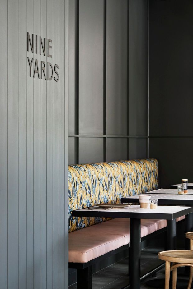 A moody, contemporary restaurant or café booth dining area. A vertically ribbed grey partition bears the words “NINE YARDS” beside a built-in banquette upholstered in a bold marbled blue, yellow, and cream pattern with pale pink seat cushions. Black tables and framing contrast with dark tiled flooring and muted grey wall panels, while small wooden stools provide seating in the foreground.