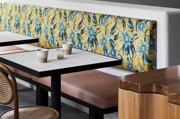 A close-up of a stylish café or dining nook featuring a long upholstered banquette with a vivid blue, yellow and green botanical pattern. Black-framed tables with pale tiled tops sit in front of the seating, accompanied by curved wooden chairs. A rounded timber counter or cabinet occupies the foreground, while textured light walls and dark flooring complete the contemporary hospitality interior.