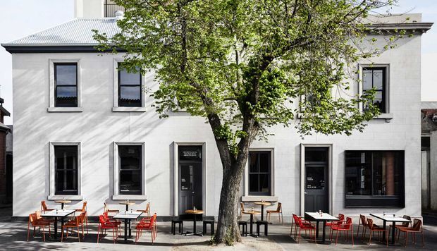 Street-facing view of a contemporary multi-storey hospitality or commercial building with a pale rendered facade, dark-framed windows and doors, a corrugated metal roof, and an outdoor cafe terrace with tables and chairs along the pavement. A mature street tree partially screens the facade.