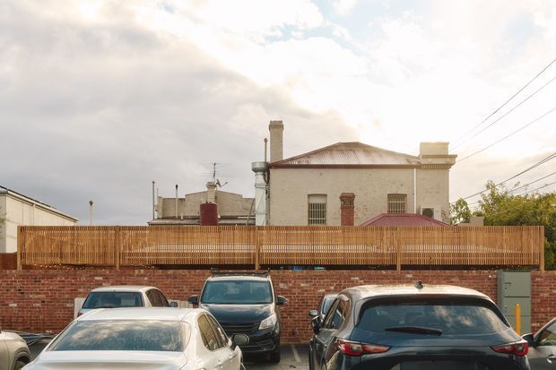 Street-level exterior view of a low-rise masonry building behind a red brick wall and timber privacy fence. The building has a pale rendered facade, hipped corrugated metal roof, chimney stacks, rooftop utility elements, and barred or louvred windows. Parked vehicles occupy the foreground, with overhead utility wires and a suburban/urban streetscape visible around the property.