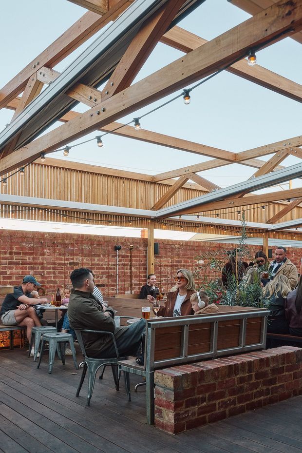 A lively outdoor dining and lounge area in a brick-walled courtyard, furnished with communal timber-and-metal seating, tables, stools, and potted planting. The space has a timber deck, exposed timber post-and-beam roof structure, translucent or open roof panels, timber slatted screening, and festoon lighting. People are gathered socially around tables, giving the venue a welcoming hospitality atmosphere.
