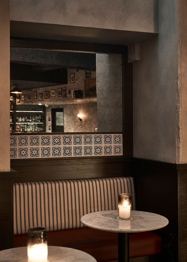 A moody, intimate restaurant or bar dining nook with a striped upholstered banquette, small round marble-topped tables, candlelight, dark wood trim, textured plaster walls, and a large mirror reflecting framed artwork and shelving. Decorative patterned tile forms a band across the lower portion of the mirror.