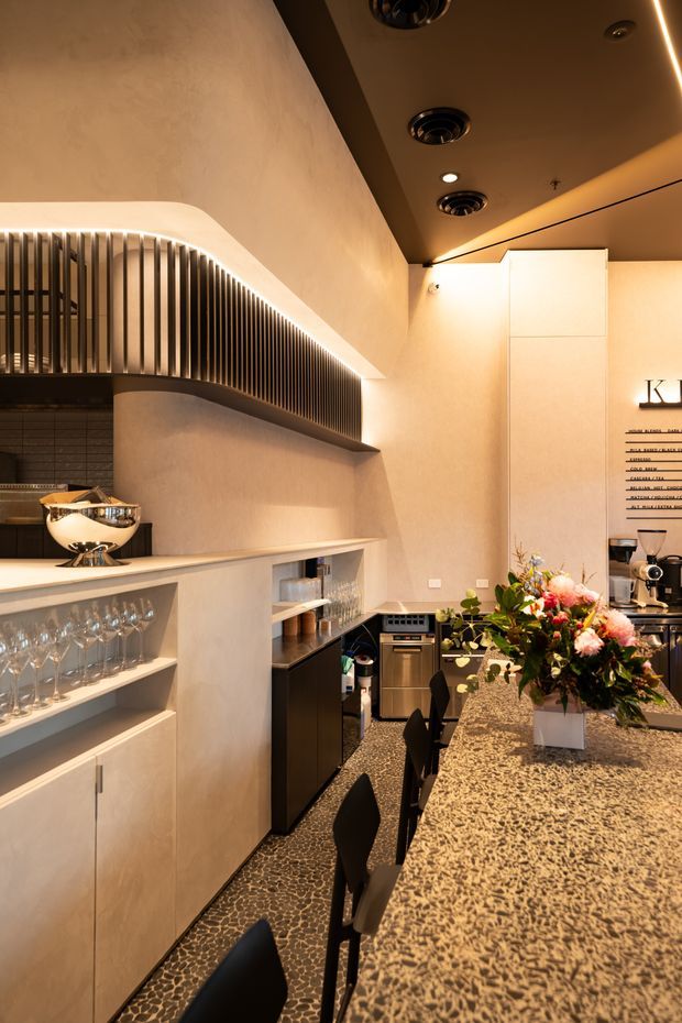 A warmly lit contemporary bar and hospitality-style kitchen interior. A long speckled stone island with dark bar stools dominates the foreground, with integrated cabinetry, glassware shelving, appliances, and a coffee or service counter along the walls. Curved plasterwork and a prominent vertical dark slatted feature with concealed lighting add architectural interest. The ceiling has recessed spotlights and linear LED lighting, while floral arrangements provide a small decorative accent.