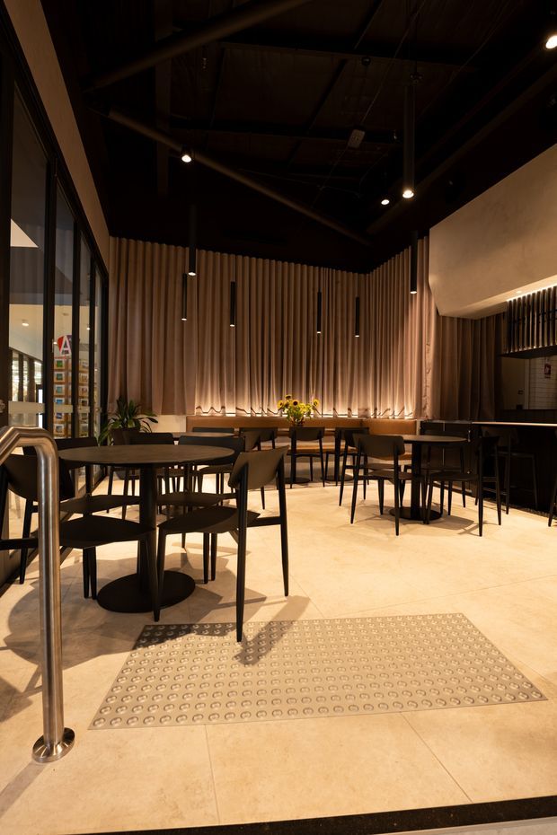 A warm, dimly lit contemporary commercial dining or café interior with numerous dark tables and chairs arranged across a pale tiled floor. The rear wall features full-height vertical timber slat panelling with integrated low-level lighting, while black exposed ceiling structure, suspended pendant lights, and dark framing create a dramatic contrast. Glazed partitions line the left side, and a tactile entry mat occupies the foreground.