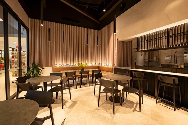 Warmly lit contemporary commercial dining and bar interior with numerous small round tables and dark chairs, a long upholstered banquette against a vertical timber slat feature wall, and a dark bar counter with stools beneath a decorative black slatted partition. Large glazed openings and retail shelving are visible at the left.