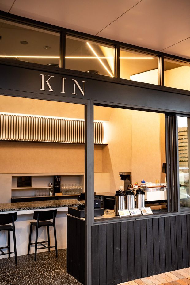 A warmly lit commercial café or bar serving area viewed through a black-framed glazed frontage. The space features a counter with beverage dispensers and coffee equipment, black bar stools, dark vertical timber-style wall panelling, a speckled dark countertop, patterned dark flooring, and an illuminated linear ceiling installation. The word “KIN” is displayed prominently on the upper frontage.