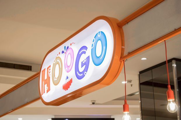 Close-up of a colourful illuminated hanging sign inside a modern commercial interior. The sign has a rounded orange frame and bright graphic lettering, suspended beneath a ceiling with exposed linear services, recessed lighting, and red pendant bulbs. Glass-fronted spaces and a mezzanine-style balustrade are visible in the background.